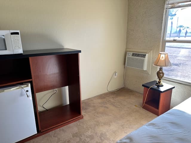 Inside a dorm room: a microwave and small refrigerator in a wood cabinet, a bedside table with a lamp, and a window.