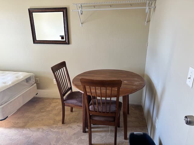 Inside a dorm room: a round wooden table with two chairs, a wall mirror and a clothes rack.