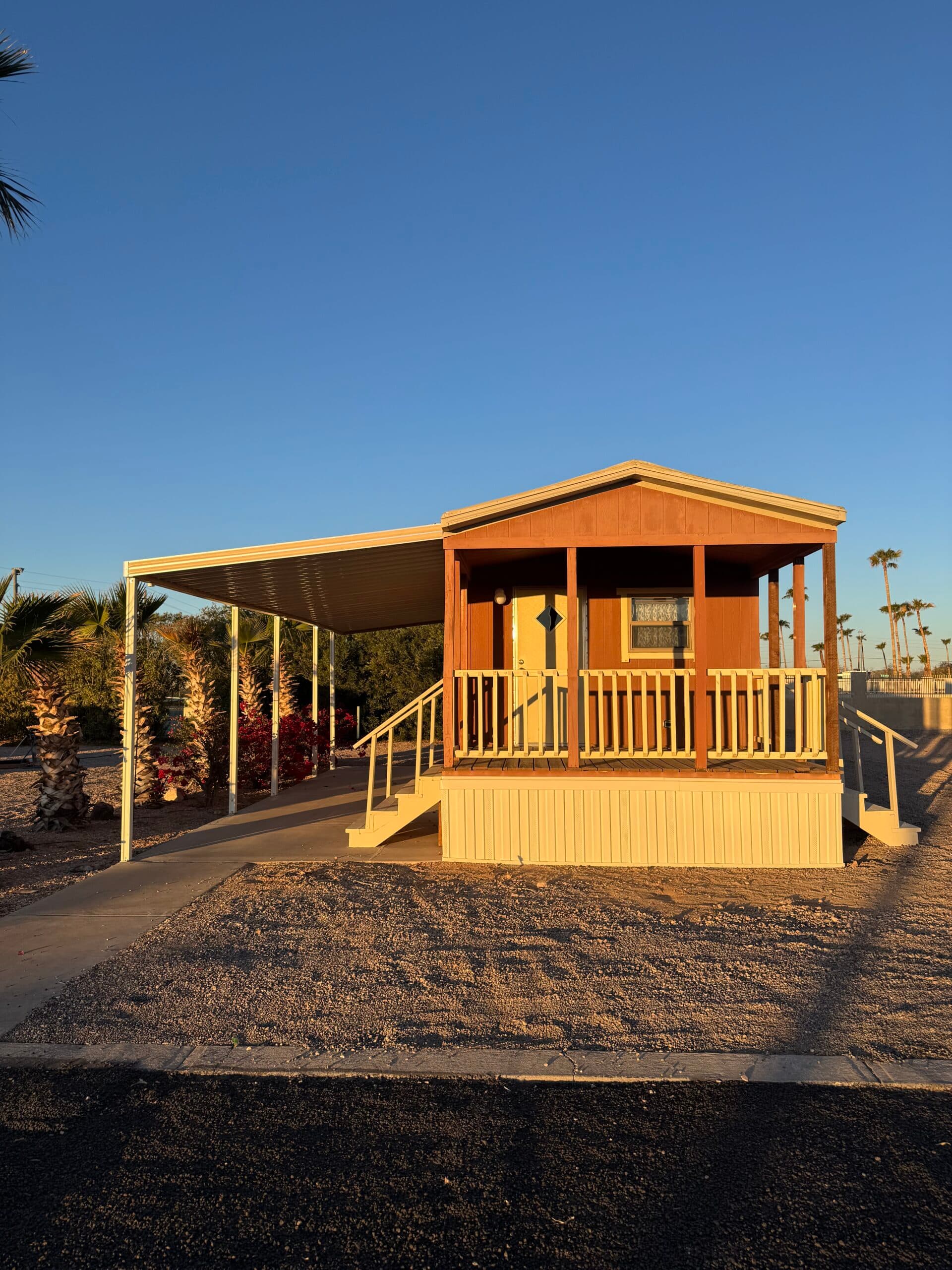 A park model with a front porch and a covered carport on a gravel lot at Saddle Mountain RV Park, with palm trees behind it.